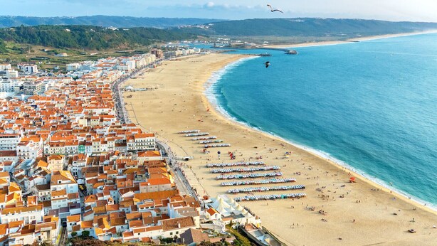 Un paysage urbain côtier avec une plage bordée de rangées de parasols et de tentes, une mer bleue et limpide, et une zone urbaine dense avec des bâtiments aux toits de tuiles rouges.