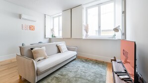 A modern living room with a sofa, a flat-screen TV, a desk with a computer, and a window with a view.