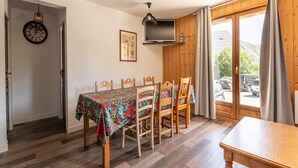 Ein Essbereich mit Tisch für vier Personen, Holzstühle, einer Wanduhr, einem flachbildfernseher über dem Tisch und Blick ins Freie durch eine Glasschiebetür.