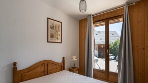 Ein Schlafzimmer mit einem hölzernen Bett, einem kleinen Nachttisch mit Lampe, einem gerahmten Bild an der Wand und einem Fenster mit Vorhängen.