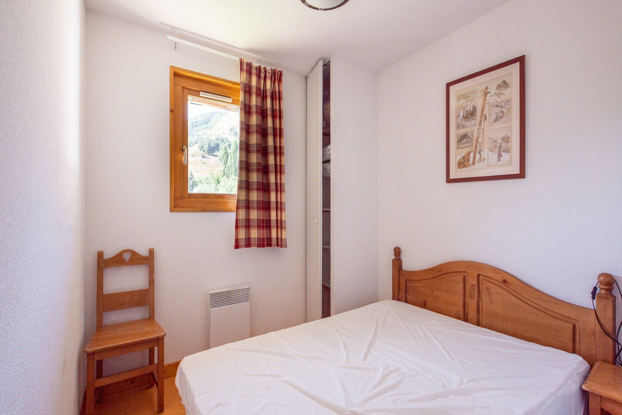 Un dormitorio con una cama de madera, una silla, un ventanal con cortina escocesa y un cuadro enmarcado en la pared.