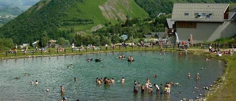 A natural pool surrounded by people, with mountains in the background and a building with a sloped roof.
