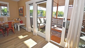 A room with a wooden floor, a dining area with a table and chairs, a sofa, and a sliding glass door leading to an outdoor area with a view of trees and a building.