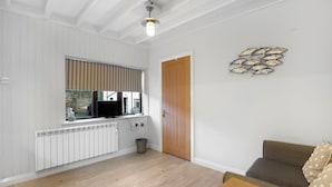 A living room with a wooden floor, a brown sofa, a wooden coffee table, a TV on a window sill, a radiator, and a door. - Minnow's Rest - North East Escapes (Seahouses)
