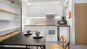 A compact kitchen with white cabinets, a black countertop, and a microwave. There is a small oven and a wooden chair. - Minnow's Rest - North East Escapes (Seahouses)