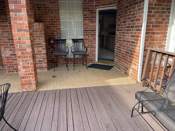 A covered patio with a brick wall, a wooden deck, two metal chairs, a fire extinguisher, and a door leading to a kitchen.