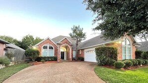 A two-story house with a brick facade, a garage, and a driveway leading to the entrance. - Charming 3-bedroom house near AT&T Stadium, Six Flag, DFW Airport (Bedford)