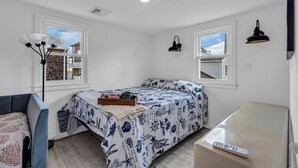 Quarto com cama, escrivaninha, luminária e duas janelas.