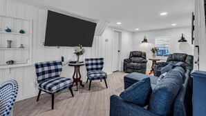 Uma sala de estar moderna com uma televisão de tela plana grande, cadeiras azulejadas e brancas, um sofá cinza e uma mesa de centro de madeira.