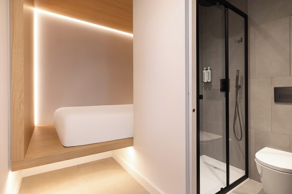 A modern bathroom with a glass shower enclosure, a toilet, and a bedside table with a mattress.