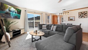 A modern living room with a sectional sofa, a flat-screen TV, a ceiling fan, and a large window offering a view of a deck. - Spacious 5-bedroom house in welcoming Manhattan (Manhattan)