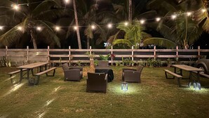 Un área de estar al aire libre con mesas y sillas, luces colgantes y palmeras.