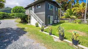 A two-story house with a covered porch, a driveway, and a fenced yard with tropical plants. - Oahu Oceanfront Getaway (Hauula)