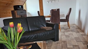 Una sala de estar moderna con un sofá de cuero negro, una mesa de centro de vidrio y un jarrón con tulipanes. Cuenta con techo de madera, un área de comedor con sillas y un reloj en la pared.