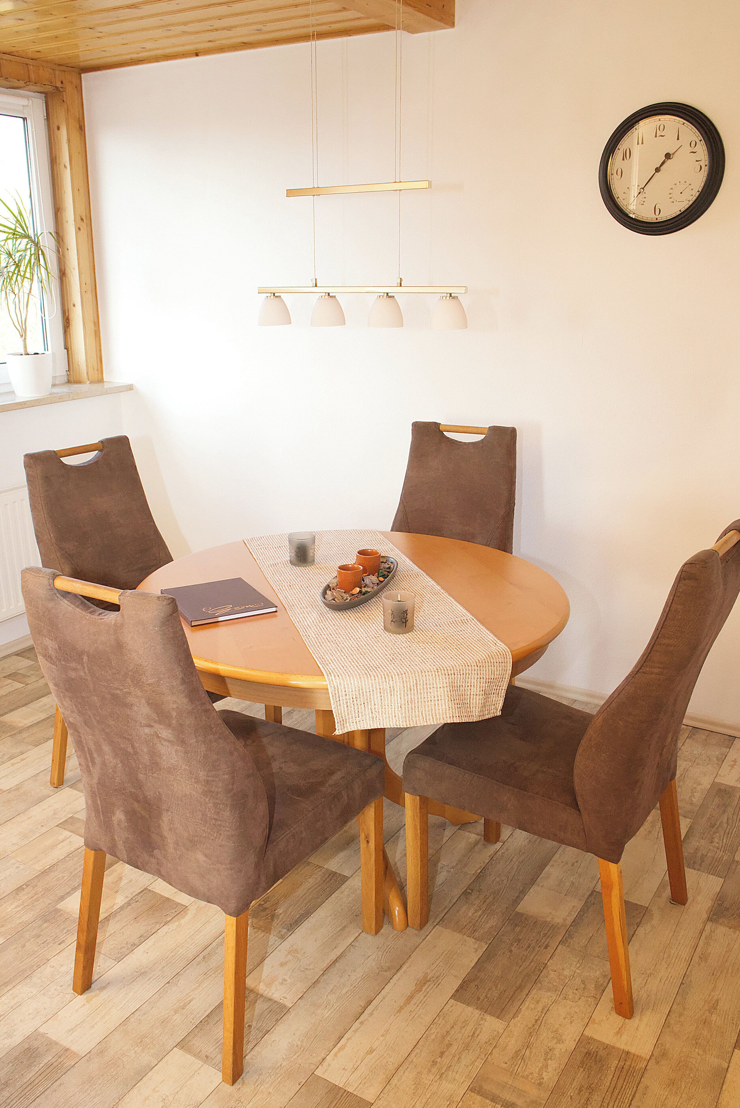 A dining area with a round wooden table, four brown upholstered chairs, a woven placemat, a small candle, and a clock on the wall.