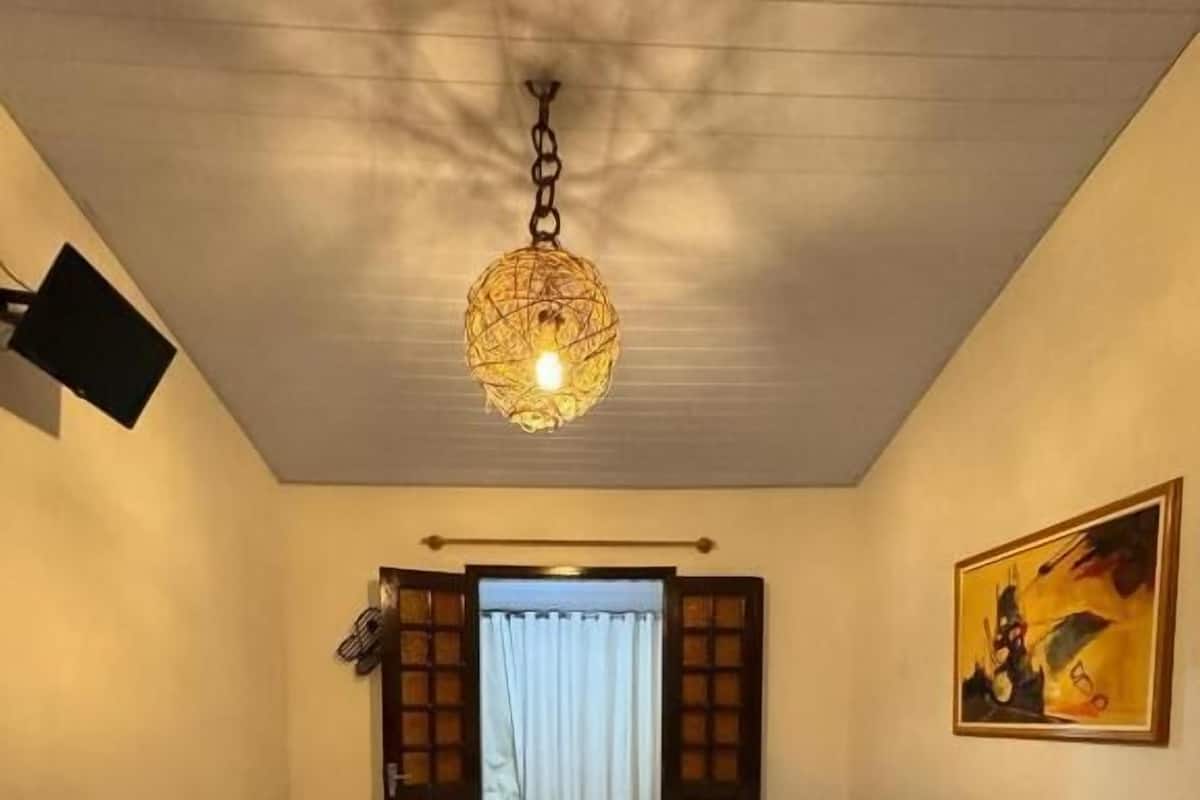 Une chambre à coucher comprenant un lit, une table de chevet, une commode, un luminaire suspendu, une fenêtre avec des rideaux et un tableau accroché au mur.