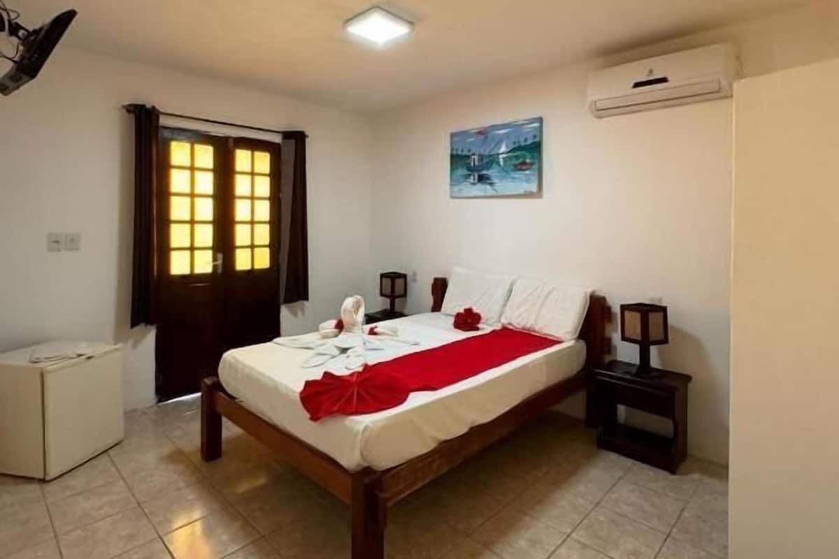 Une chambre à coucher comprenant un lit, une table de chevet, un climatiseur fixé au mur, une fenêtre avec des rideaux et un tableau accroché au mur.