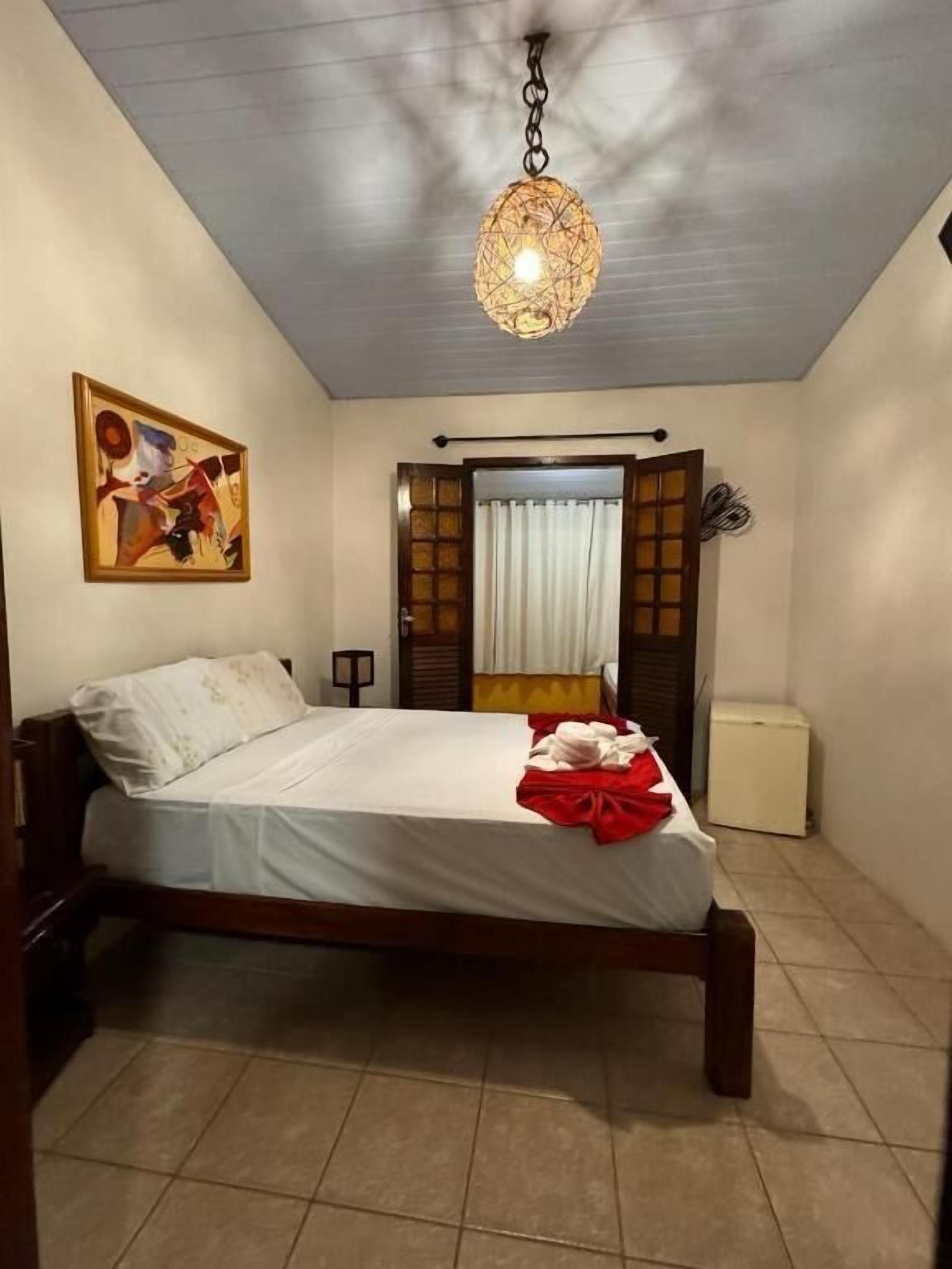 Een slaapkamer met een bed, een schilderij aan de muur, een plafondlamp en een raam met gordijnen.