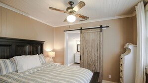 Un dormitorio con cama, ventilador de techo, mesita de noche con lámpara, armario de madera y baño visible a través de un vano.