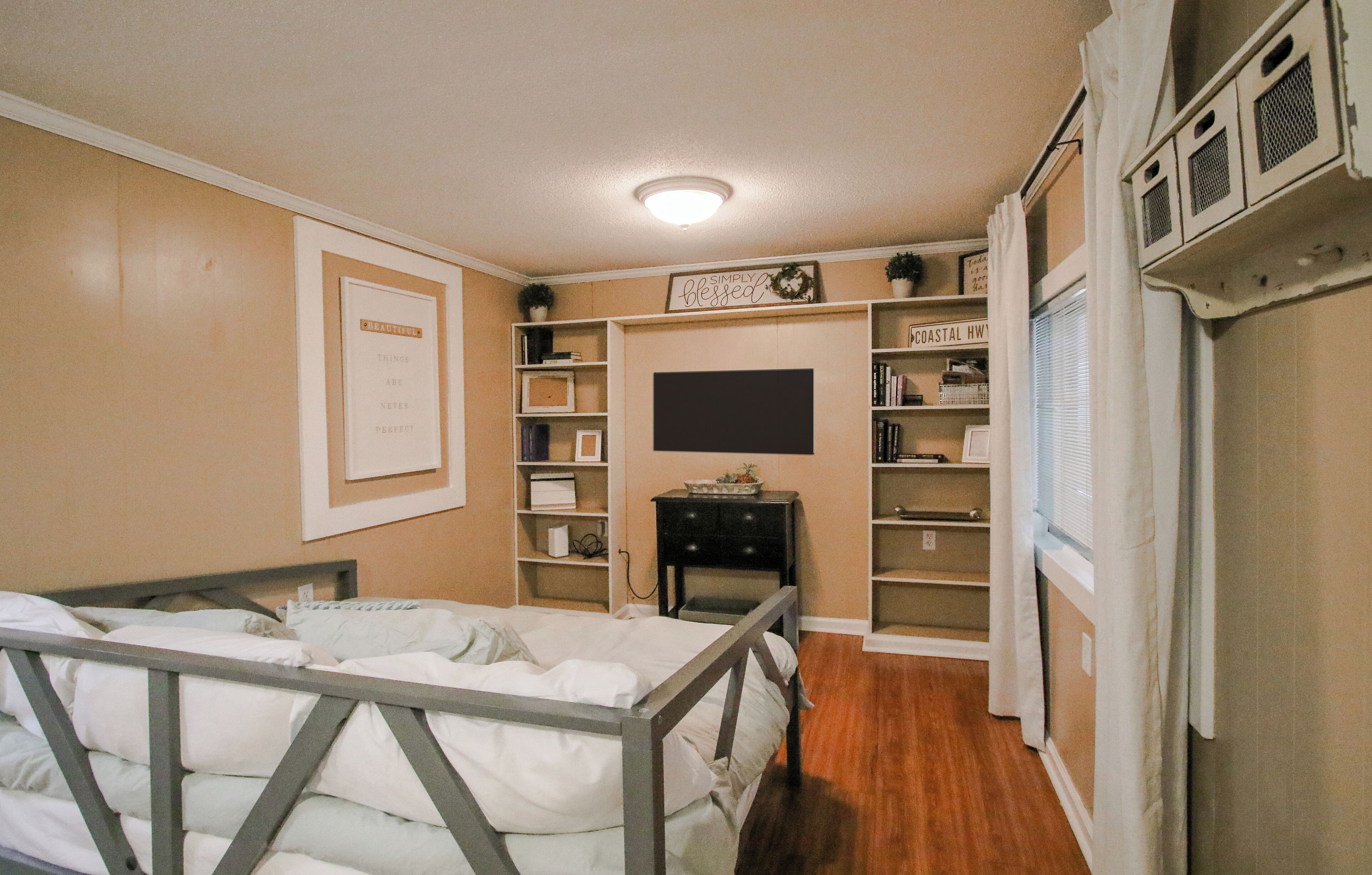 Une chambre à coucher comprenant un lit, des étagères, un téléviseur à écran plat et une fenêtre avec des stores.