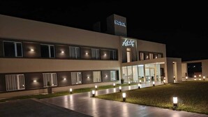 Exterior moderno de un hotel por la noche, con una entrada bien iluminada, un letrero visible y un área pavimentada con pequeñas luces.