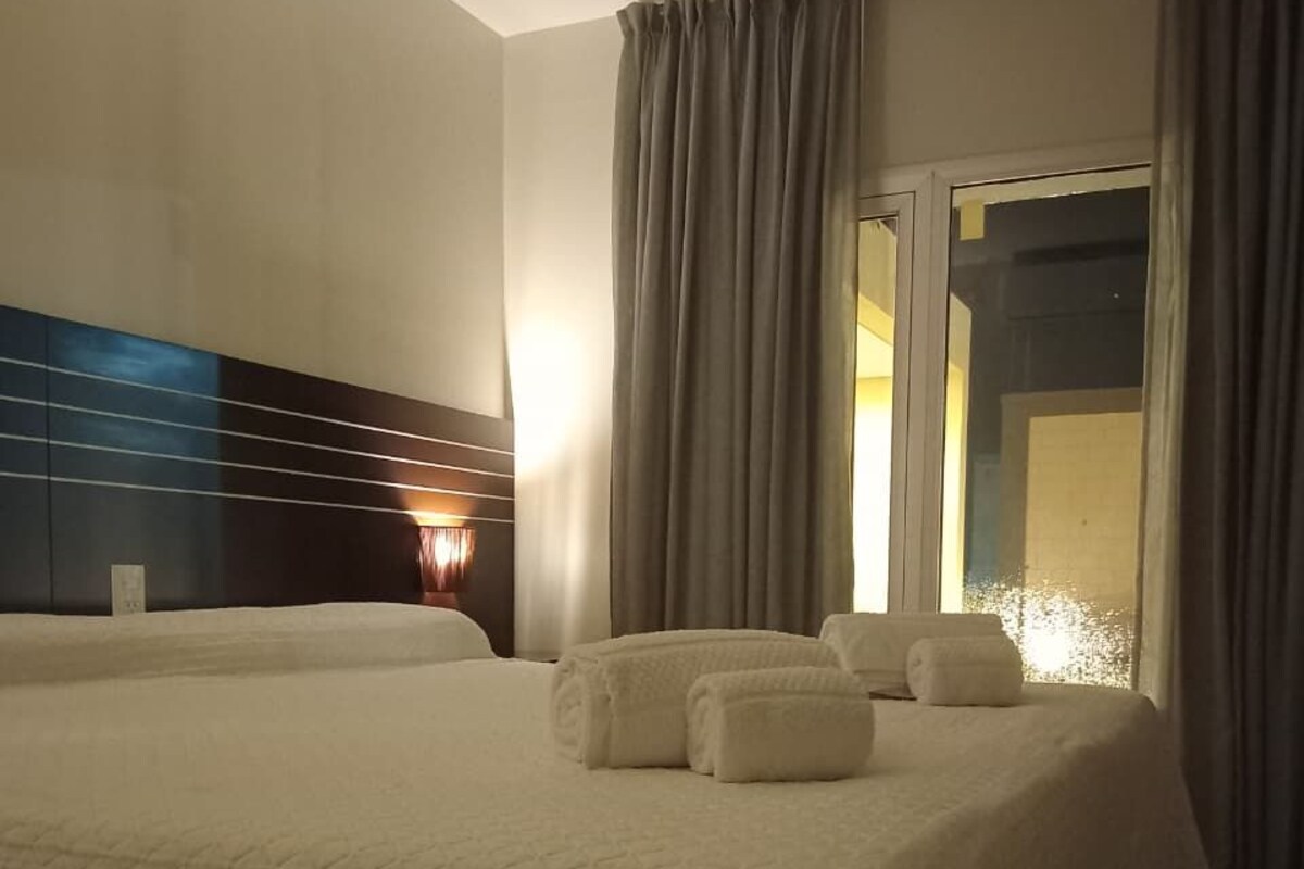 Una habitación de hotel con una cama grande, cabecera con líneas horizontales, una mesita de noche y una ventana con cortinas.