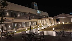Exterior de un hotel por la noche, con mesas al aire libre y senderos iluminados.