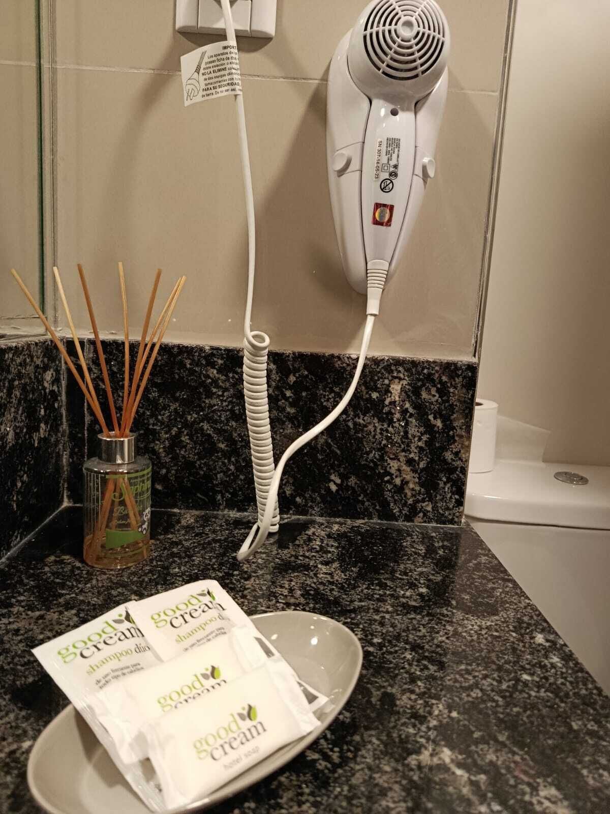 A bathroom with a hairdryer, a bottle of reed diffuser, and a bowl of Good Cream shampoo.
