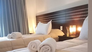 Una habitación de hotel con dos camas, cada una con cabecero y almohadas. Hay toallas enrolladas sobre las camas y una mesita de noche con una lámpara y una botella de agua.