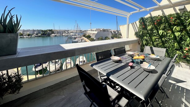 Une terrasse avec une table et des chaises, donnant sur un port de plaisance où sont amarrés des bateaux.