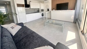 Un salon moderne avec un canapé gris, une table basse en verre et un coin cuisine doté de meubles blancs et d’un téléviseur à écran plat.