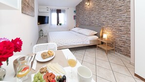A modern bedroom with a stone wall, a bed with white bedding, a bedside table with a lamp, and a breakfast setup with fruits, cheese, and a drink. - Charming studio with sea view, bikes, baby crib, barbecue (Crikvenica)