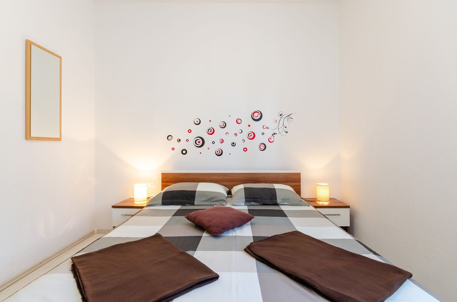 Een net opgemaakt bed met een geruit sprei en twee bruine handdoeken, een muur met een decoratief rond patroon, en twee bedlampjes.