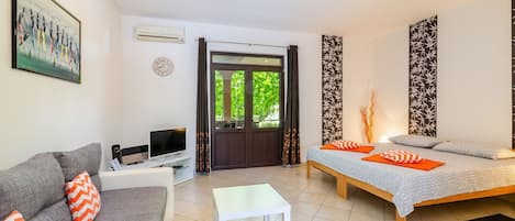 Un salon moderne avec un canapé, un téléviseur, un lit avec une literie à motifs et une grande fenêtre avec des rideaux.