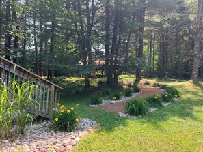 A forested area with a wooden deck, a gravel path, and various plants.