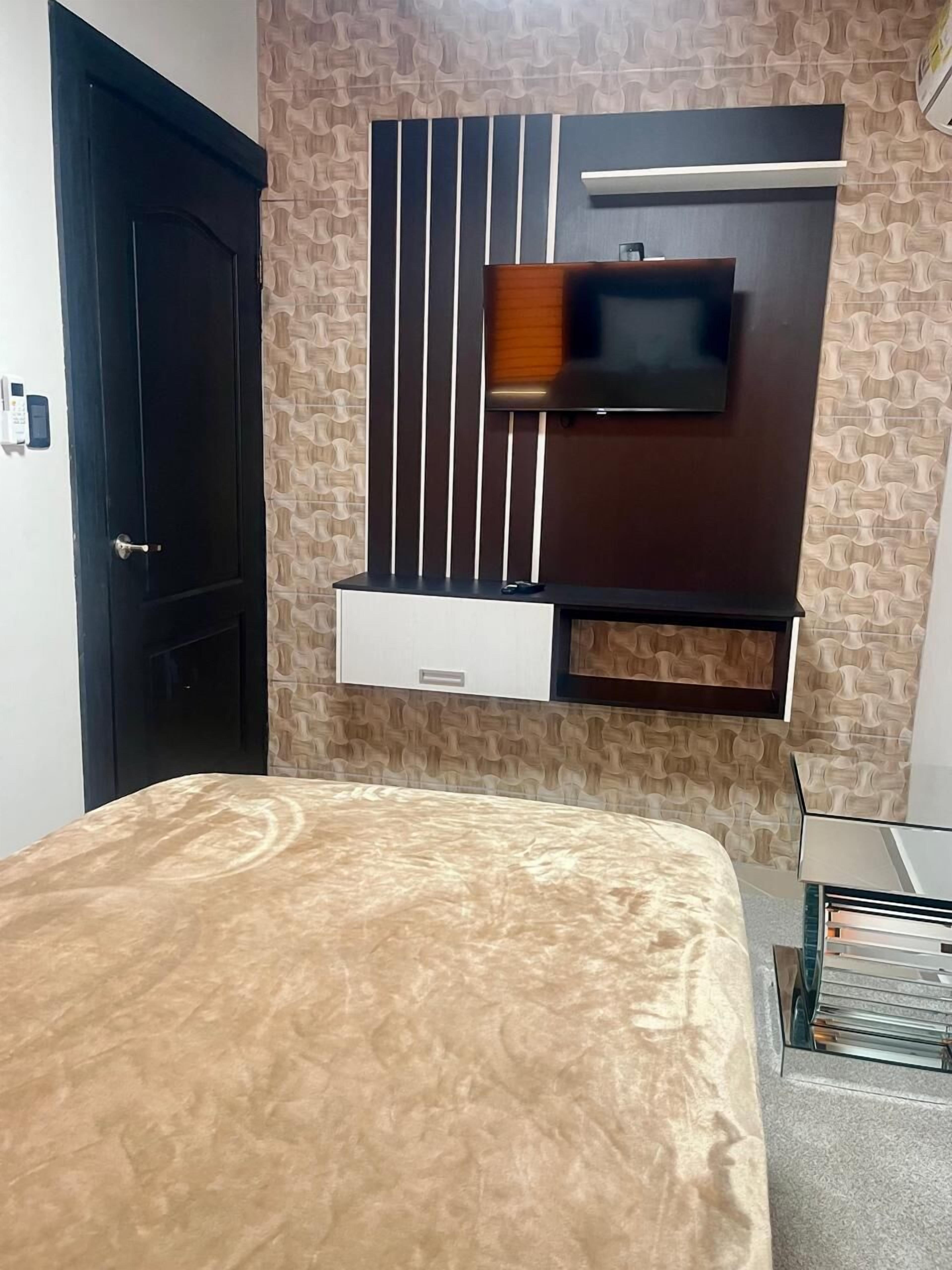 Ein Schlafzimmer mit einem Bett, einem an der Wand montierten Fernseher, einer Tür und einem kleinen Schrank.