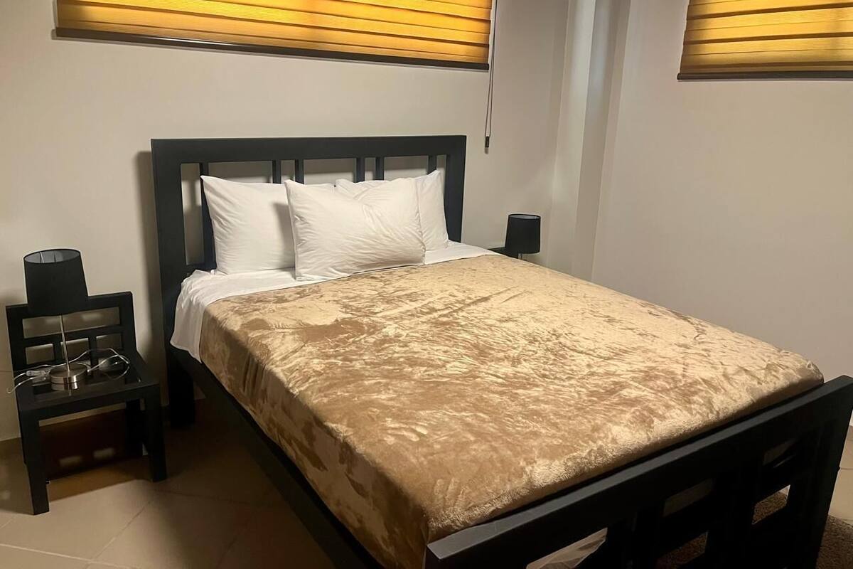 Un lit bien fait, recouvert d’une couvre-lit à motifs dorés, avec une tête de lit en bois foncé et deux lampes de chevet.