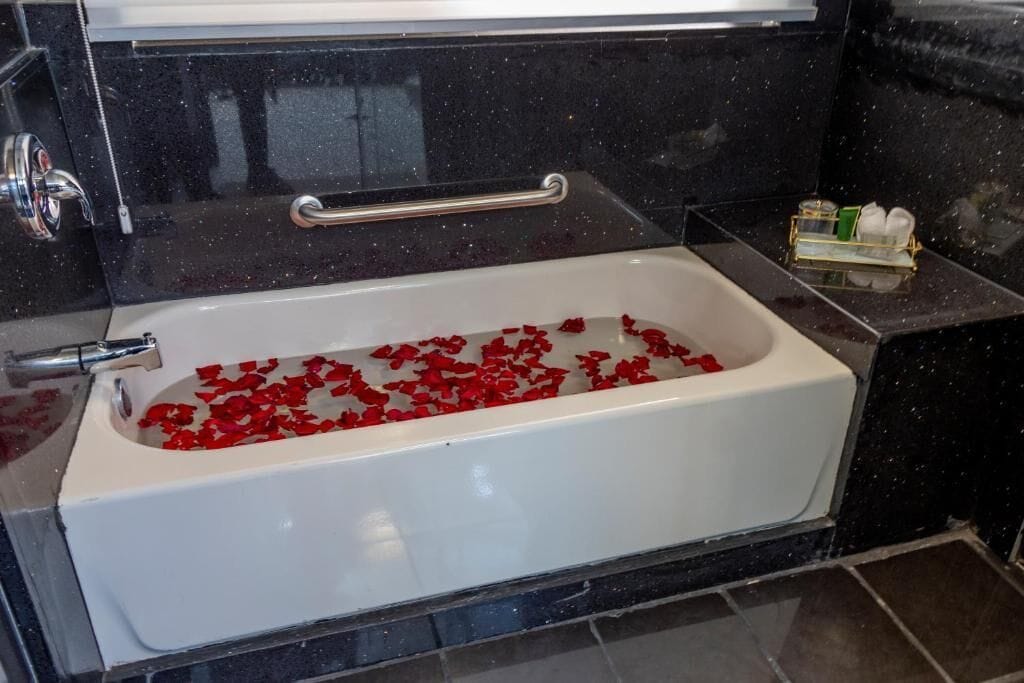 A bathtub filled with rose petals, a towel rack, and toiletries on a black countertop.