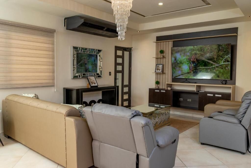 Una sala de estar moderna con un sofá color beige, sillones grises, un televisor de pantalla plana y un espejo decorativo.