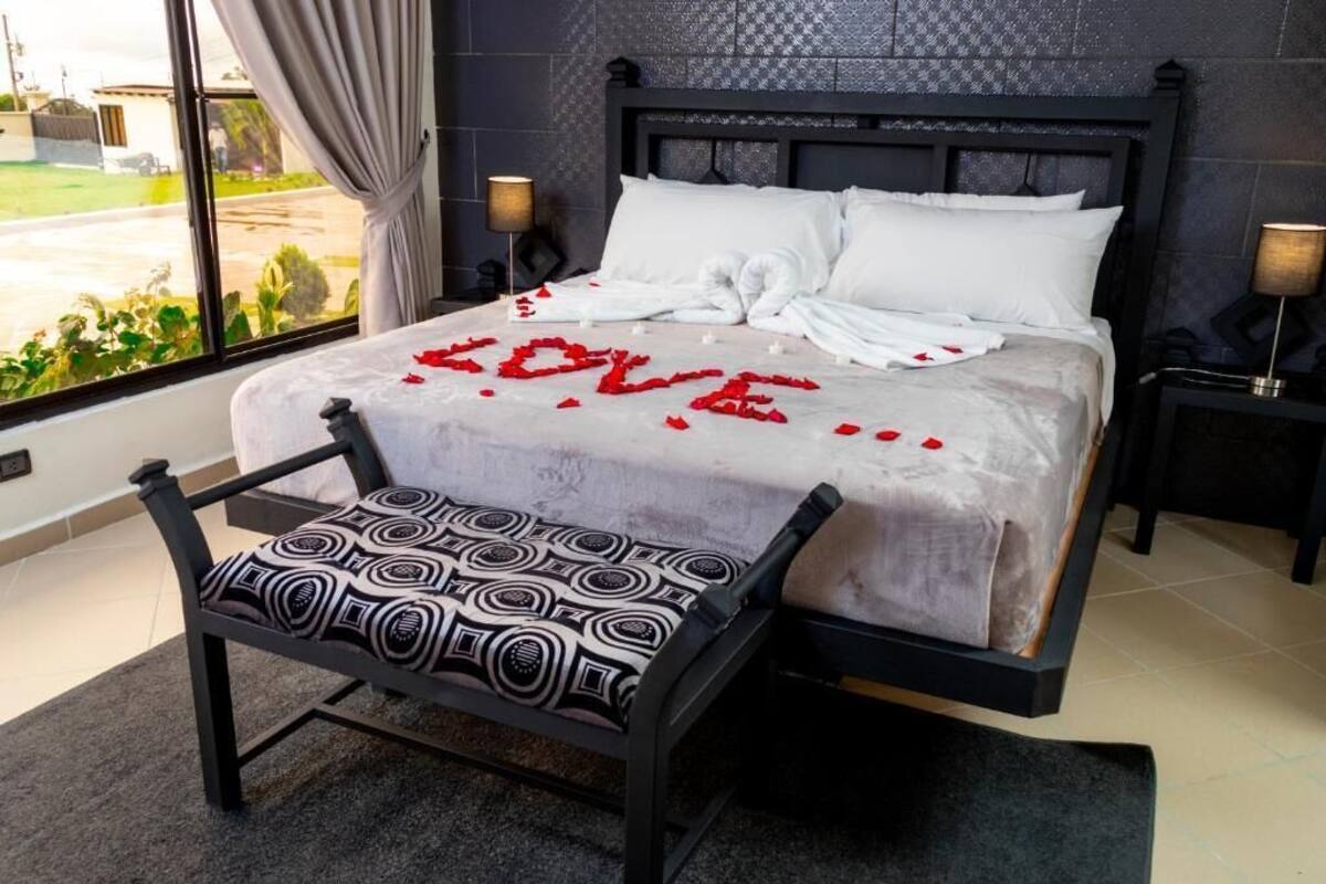 Un lit parfaitement fait, avec des draps blancs, et une composition en forme de cœur, faite de pétales de roses, qui épelle le mot « AMOUR » sur la couverture.