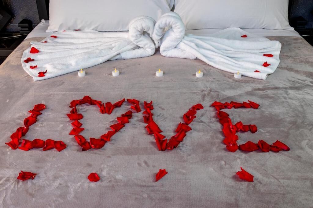 Un letto con petali di rosa rossa disposti a forma di cuore e la scritta 'LOVE', insieme a un paio di asciugamani bianchi piegati a cuore.