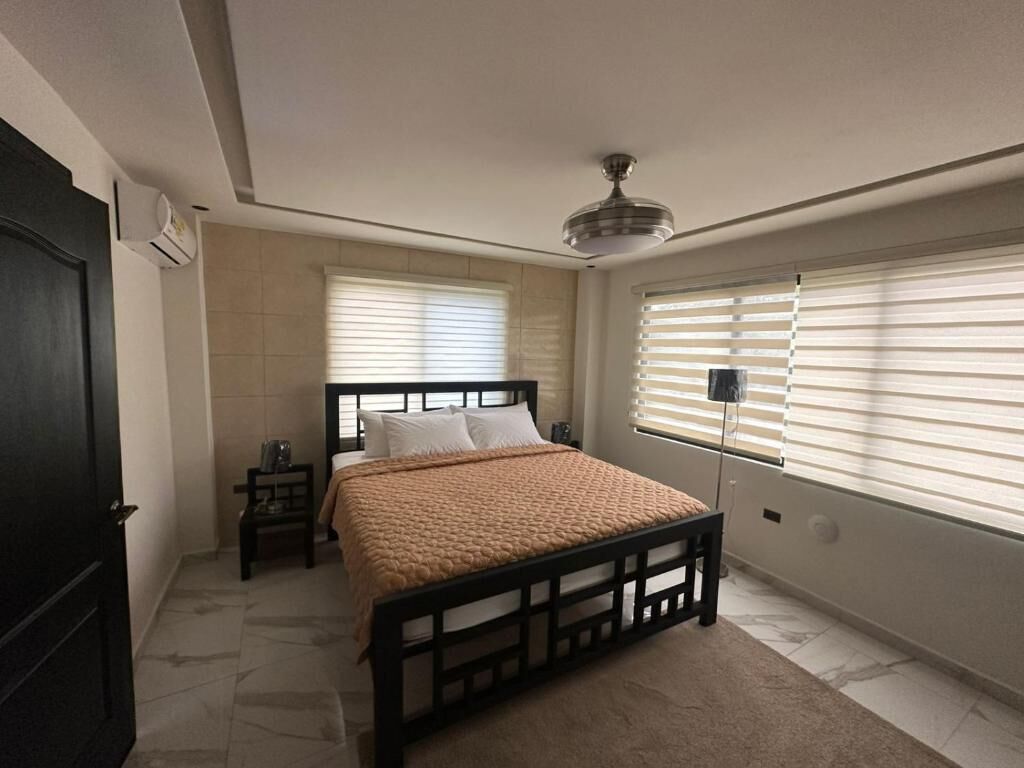 Ein Schlafzimmer mit einem Bett, einem Nachttisch, einem Deckenventilator und einem Fenster mit Jalousien.