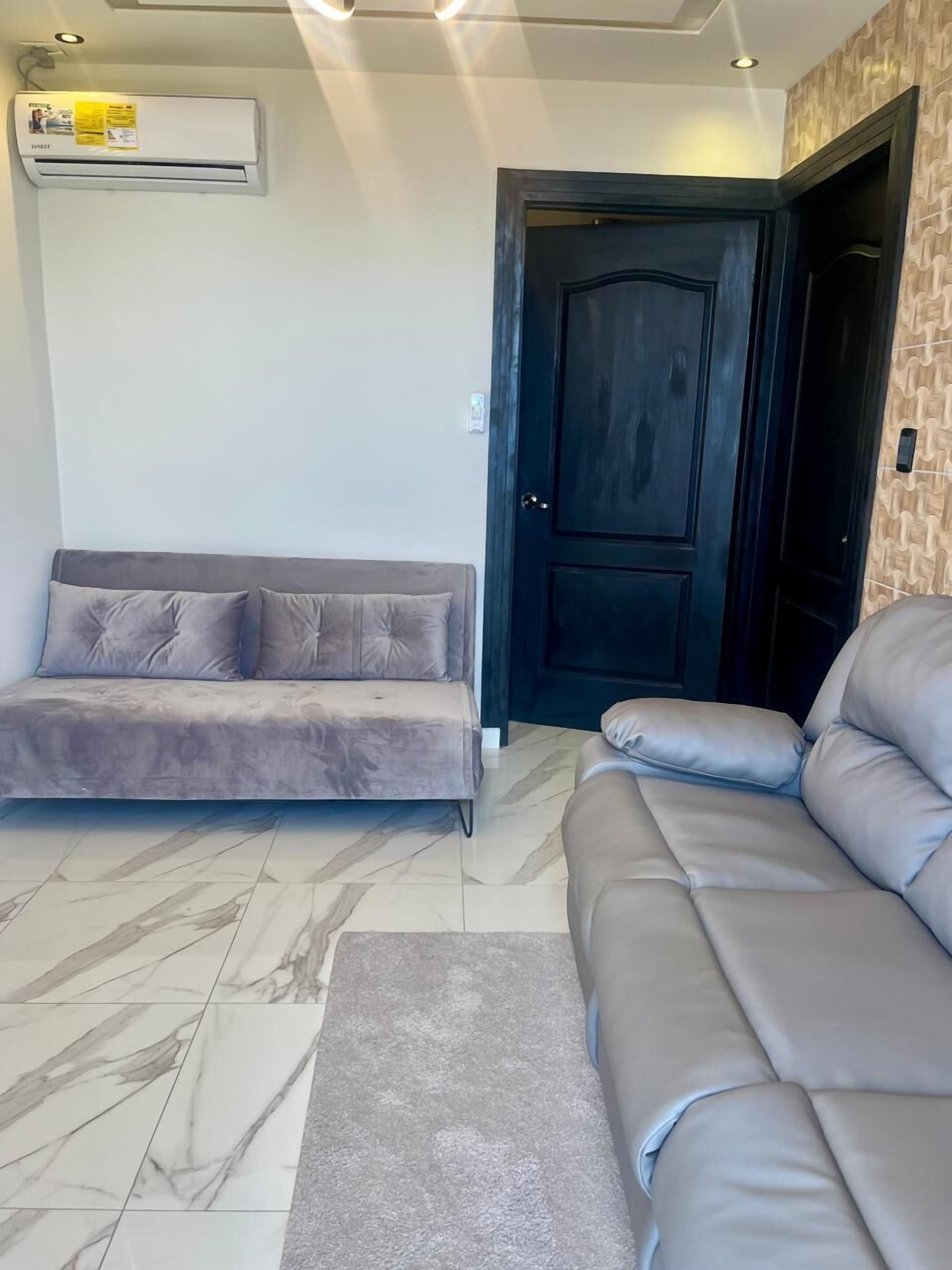 Una sala de estar con un sofá gris, un sofá seccional gris más grande, una puerta de madera oscura y una unidad de aire acondicionado en la pared.