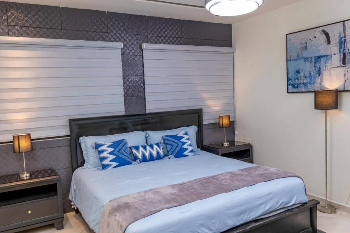 Une chambre à coucher comprenant un lit, deux tables de chevet, une œuvre d’art accrochée au mur et une fenêtre avec des stores.