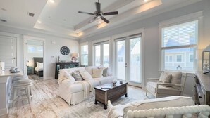 A spacious living room with a white sofa, a coffee table, and a chair. There is a ceiling fan and large windows providing natural light.