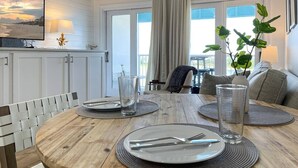 Un comedor con una mesa de madera dispuesta para cuatro personas, un televisor montado en la pared y vistas al exterior a través de amplias ventanas.