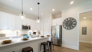 Cocina moderna con gabinetes blancos, refrigerador de acero inoxidable, un reloj grande y tres taburetes.