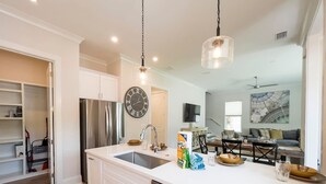 Una cocina moderna con armarios blancos, un refrigerador de acero inoxidable, un lavavajillas y una isla grande con un fregadero. Hay un reloj en la pared y un ventilador de techo al fondo.