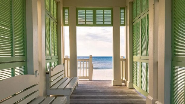Una terraza de madera que da a una playa, con contraventanas verdes y un banco.