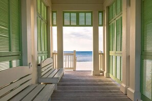 Una terraza de madera que da a una playa, con contraventanas verdes y un banco.